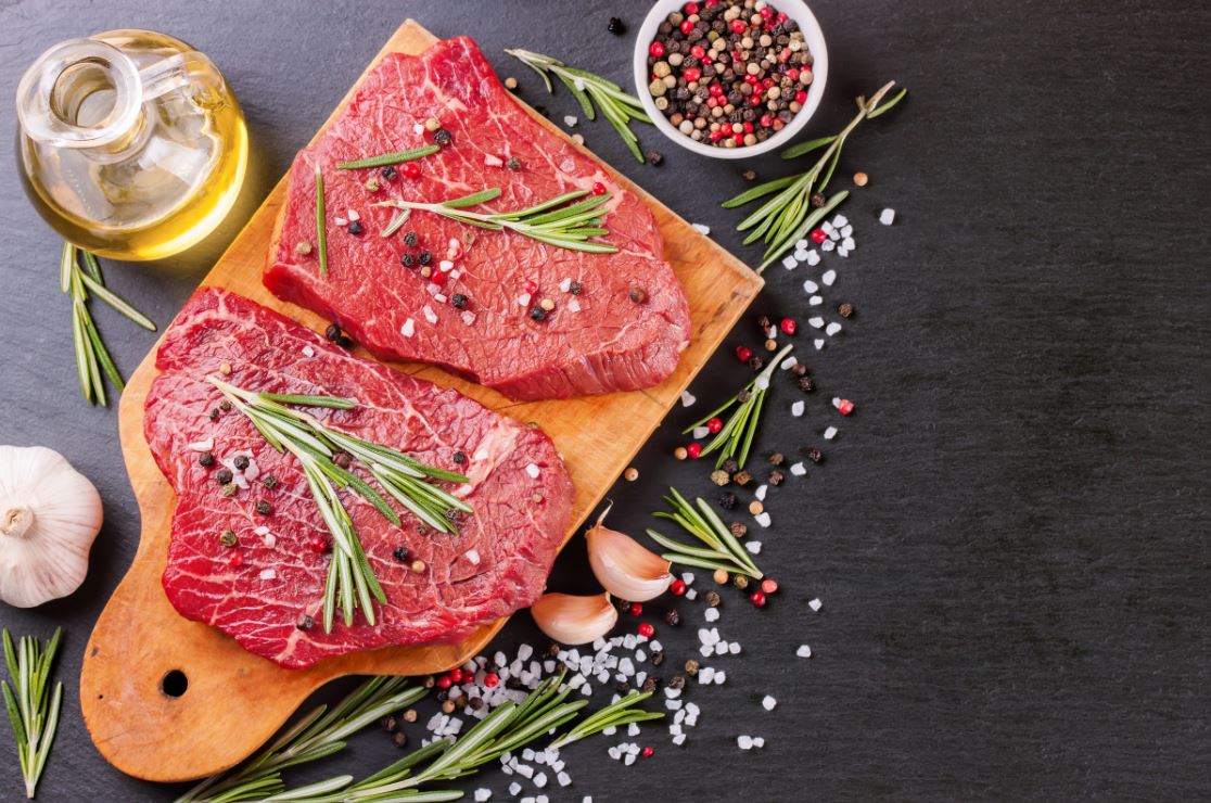 Raw steak on a wooden chopping board surrounded by herbs and spices